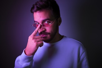 Male model adjusting his grey-rimmed eyeglasses, with a soft purple light in the background.
