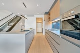 A modern kitchen with sleek design elements, featuring a long island with an integrated sink and a black faucet. The kitchen cabinets are minimalist with flat surfaces, and a built-in oven is visible. A staircase with a glass railing is on the left side, leading to an upper floor. Light wood flooring and cabinets add warmth to the bright space.