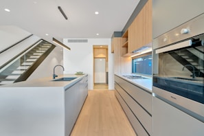 A modern kitchen with sleek design elements, featuring a long island with an integrated sink and a black faucet. The kitchen cabinets are minimalist with flat surfaces, and a built-in oven is visible. A staircase with a glass railing is on the left side, leading to an upper floor. Light wood flooring and cabinets add warmth to the bright space.