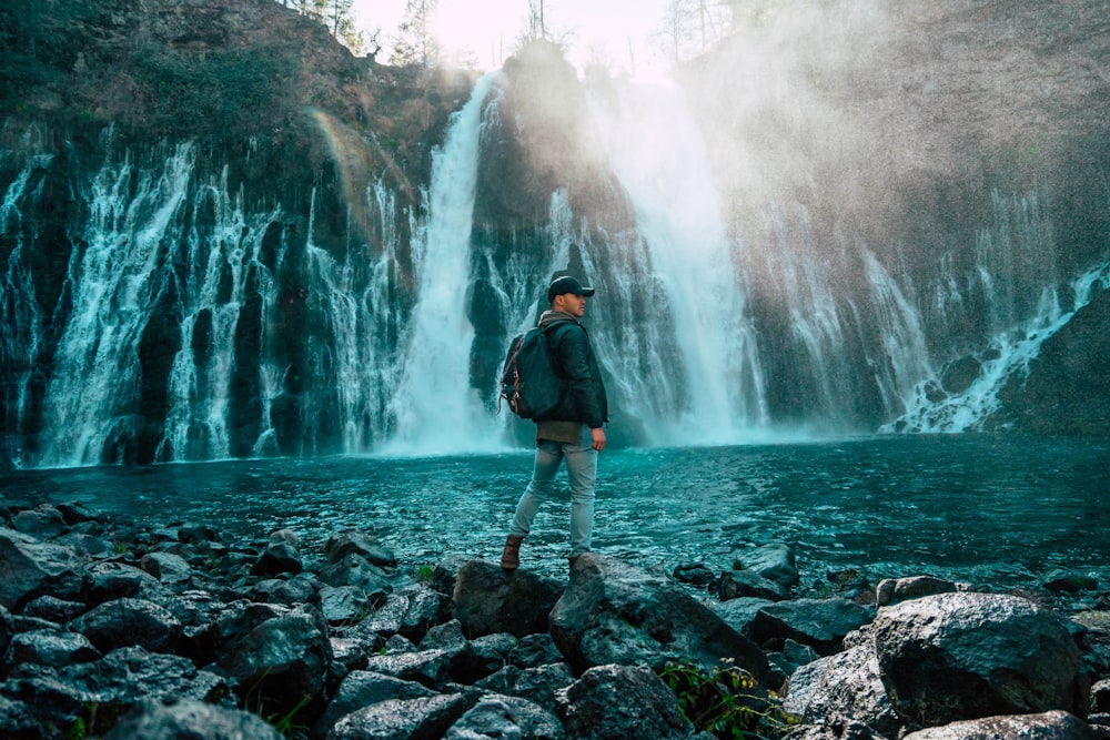 man in black jacket standing on rocky shore near waterfalls during daytime