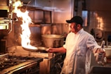 A chef in a commercial kitchen is flambéing a dish in a frying pan. The flame rises dramatically as the chef, wearing a white coat and black cap, holds a bottle of liquor in his other hand. The kitchen setting includes a stove with multiple burners and a large pot in the background.
