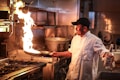 A chef in a commercial kitchen is flamb&eacute;ing a dish in a frying pan. The flame rises dramatically as the chef, wearing a white coat and black cap, holds a bottle of liquor in his other hand. The kitchen setting includes a stove with multiple burners and a large pot in the background.