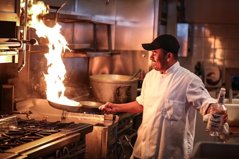 A chef in a commercial kitchen is flambéing a dish in a frying pan. The flame rises dramatically as the chef, wearing a white coat and black cap, holds a bottle of liquor in his other hand. The kitchen setting includes a stove with multiple burners and a large pot in the background.