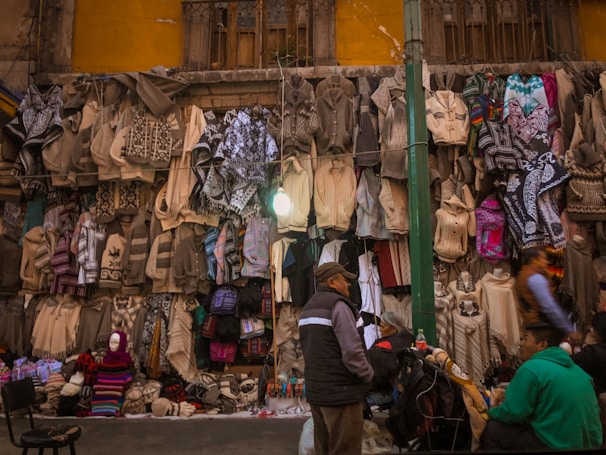 A street market scene with a variety of woven textiles hanging on a wall. Sweaters and ponchos in shades of beige, gray, and some colorful patterns are displayed. People are browsing the textiles, with a few seated near the stall. The background includes an old, weathered building facade with a window and a light bulb hanging above the display.