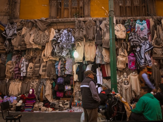 A street market scene with a variety of woven textiles hanging on a wall. Sweaters and ponchos in shades of beige, gray, and some colorful patterns are displayed. People are browsing the textiles, with a few seated near the stall. The background includes an old, weathered building facade with a window and a light bulb hanging above the display.