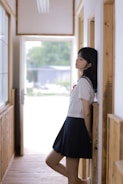 A person in a school uniform leans against a wall in a hallway. The setting is bright, with natural light coming through large windows. Wooden elements are present in the architecture, giving a warm and serene feel.