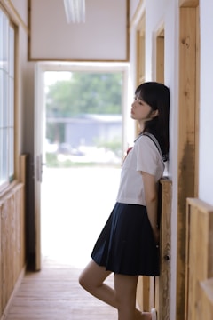 A person in a school uniform leans against a wall in a hallway. The setting is bright, with natural light coming through large windows. Wooden elements are present in the architecture, giving a warm and serene feel.