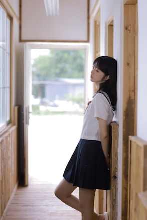 A person in a school uniform leans against a wall in a hallway. The setting is bright, with natural light coming through large windows. Wooden elements are present in the architecture, giving a warm and serene feel.