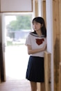 A young woman is standing in a hallway, partially leaning against a wooden wall. She is wearing a school uniform, consisting of a white blouse with a sailor-style collar and a red bow, and a dark pleated skirt. The background is softly lit with a window that shows some greenery outside.