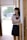 A young woman is standing in a hallway, partially leaning against a wooden wall. She is wearing a school uniform, consisting of a white blouse with a sailor-style collar and a red bow, and a dark pleated skirt. The background is softly lit with a window that shows some greenery outside.