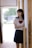A young woman is standing in a hallway, partially leaning against a wooden wall. She is wearing a school uniform, consisting of a white blouse with a sailor-style collar and a red bow, and a dark pleated skirt. The background is softly lit with a window that shows some greenery outside.