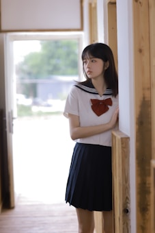 A young woman is standing in a hallway, partially leaning against a wooden wall. She is wearing a school uniform, consisting of a white blouse with a sailor-style collar and a red bow, and a dark pleated skirt. The background is softly lit with a window that shows some greenery outside.