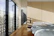 A modern bedroom with a minimalist design features a bed with white pillows and a gray duvet against a wooden accent wall. Sunlight streams in through large windows with a perforated design, creating patterns of light and shadow on the wooden floor.