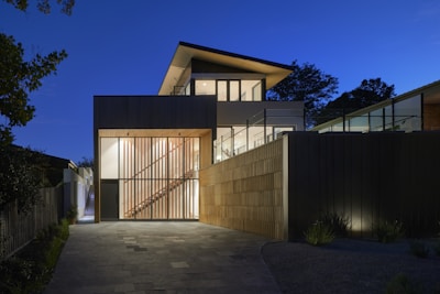 A modern two-story house with large glass windows and a wooden facade is illuminated during twilight. The architecture includes clean lines and geometric shapes, with a combination of concrete and wood materials. Soft outdoor lighting accentuates the structure's features, while the sky is clear and deep blue, adding to the tranquil ambiance.