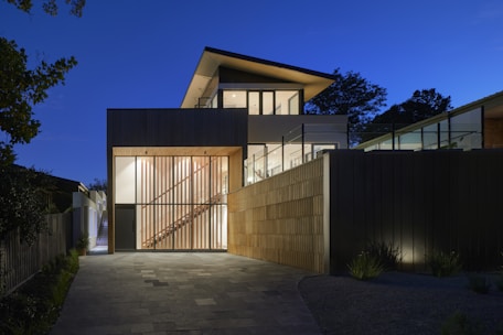 A modern two-story house with large glass windows and a wooden facade is illuminated during twilight. The architecture includes clean lines and geometric shapes, with a combination of concrete and wood materials. Soft outdoor lighting accentuates the structure's features, while the sky is clear and deep blue, adding to the tranquil ambiance.