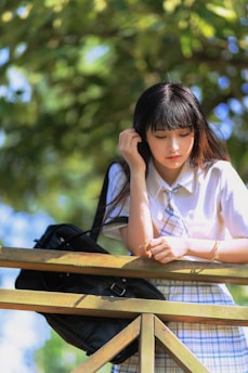 A young person with long dark hair stands pensively by a wooden railing, holding a small twig. They are wearing a white shirt with a plaid skirt and tie, suggesting a school uniform. Sunlight filters through the leafy green tree above, creating a serene and thoughtful atmosphere.