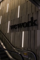A dimly lit modern interior with an escalator and a wall featuring vertical light strips. The word 'wework' is prominently displayed on the wall in bold letters. The setting appears corporate or industrial with a sleek and minimalist design.