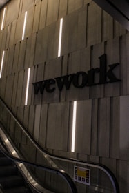 A dimly lit modern interior with an escalator and a wall featuring vertical light strips. The word 'wework' is prominently displayed on the wall in bold letters. The setting appears corporate or industrial with a sleek and minimalist design.