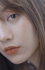 Close-up of a person's face focusing on the eye and lips, featuring natural makeup with brown lipstick. The skin appears smooth and the hair is brown, slightly covering part of the face.