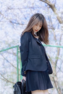 A person in a dark navy school uniform with pleated skirt, standing outside with a background of light pastel colors from blossoming flowers. The individual is holding a black bag, and appears to be looking down in a contemplative manner.