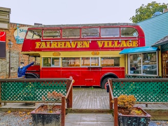 An old-fashioned red double-decker bus is situated in a quaint village setting. The words 'Fairhaven Village' are prominently displayed across the side of the bus. A mannequin with a blue jacket is posed as if boarding the bus. Surrounding the bus is a small wooden deck enclosed by green lattice fencing and overgrown planters. The scene is completed with nearby rustic buildings, contributing to a nostalgic, vintage ambiance.