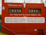 A red brick wall with vintage typography displays the history of ownership of the D.P. Mason Drug Co., established in 1889. Names and years of ownership are listed in white text on a decorative panel. A small sign for a Lutheran church is also visible.