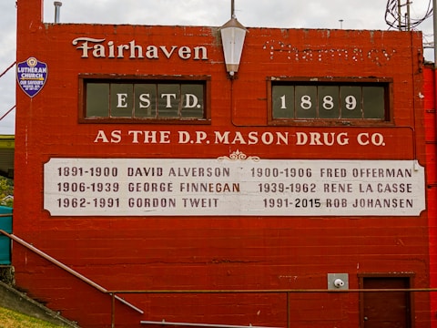 A red brick wall with vintage typography displays the history of ownership of the D.P. Mason Drug Co., established in 1889. Names and years of ownership are listed in white text on a decorative panel. A small sign for a Lutheran church is also visible.