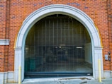 An arched garage entrance with a closed metal security gate. The building is made of red brick with white stone accents around the archway. The area appears to lead to an underground or enclosed space with minimal lighting.