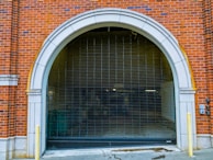 An arched garage entrance with a closed metal security gate. The building is made of red brick with white stone accents around the archway. The area appears to lead to an underground or enclosed space with minimal lighting.