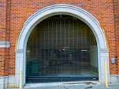 An arched garage entrance with a closed metal security gate. The building is made of red brick with white stone accents around the archway. The area appears to lead to an underground or enclosed space with minimal lighting.