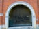 An arched garage entrance with a closed metal security gate. The building is made of red brick with white stone accents around the archway. The area appears to lead to an underground or enclosed space with minimal lighting.