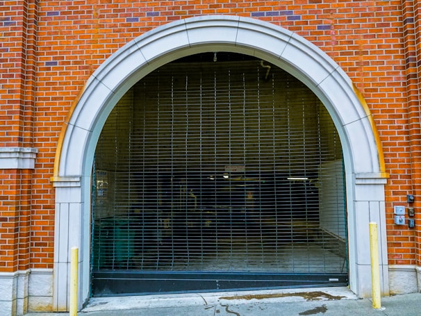 An arched garage entrance with a closed metal security gate. The building is made of red brick with white stone accents around the archway. The area appears to lead to an underground or enclosed space with minimal lighting.