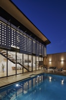 A modern architectural home featuring large glass windows and a unique perforated facade, with a view of the interior including a contemporary staircase and dining area. The exterior features a wooden deck and a blue-tiled swimming pool, with cozy outdoor seating. The scene is set during dusk, enhancing the warm lighting and reflections in the pool.