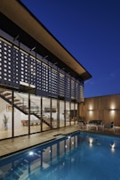 A modern architectural home featuring large glass windows and a unique perforated facade, with a view of the interior including a contemporary staircase and dining area. The exterior features a wooden deck and a blue-tiled swimming pool, with cozy outdoor seating. The scene is set during dusk, enhancing the warm lighting and reflections in the pool.