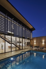 A modern architectural home featuring large glass windows and a unique perforated facade, with a view of the interior including a contemporary staircase and dining area. The exterior features a wooden deck and a blue-tiled swimming pool, with cozy outdoor seating. The scene is set during dusk, enhancing the warm lighting and reflections in the pool.