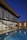 A modern architectural home featuring large glass windows and a unique perforated facade, with a view of the interior including a contemporary staircase and dining area. The exterior features a wooden deck and a blue-tiled swimming pool, with cozy outdoor seating. The scene is set during dusk, enhancing the warm lighting and reflections in the pool.
