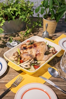 A roasted chicken is served in a yellow baking dish, garnished with mushrooms, cherry tomatoes, and fresh herbs. The dish is set on a rustic wooden table with a vibrant yellow napkin, accompanied by a fork, knife, and two plates. Two potted plants are placed in the background, adding a touch of greenery to the setting.