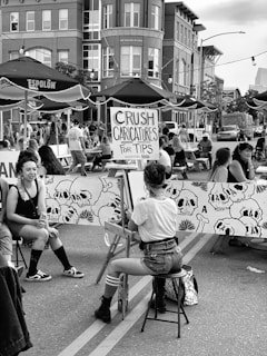A street artist is drawing caricatures for tips at an outdoor event. The setting features tall buildings and umbrellas with branded signage. Several people are seated and gathered around, creating a lively atmosphere. A prominent sign advertising caricatures is visible, and a mural of skulls is part of the backdrop.
