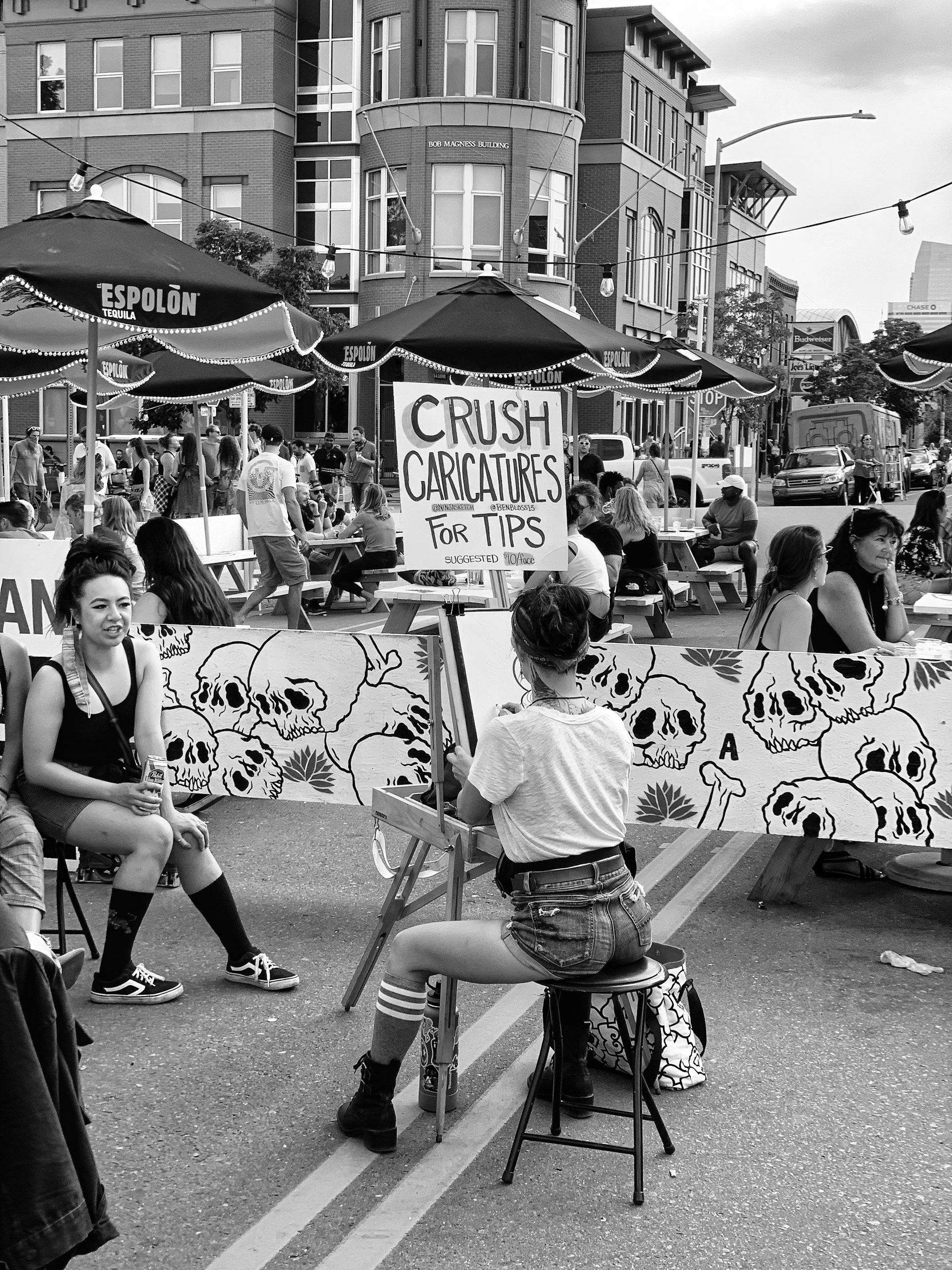 A colorful festival scene featuring a crowd enjoying live caricature drawings, with John Valcas sketching on the spot.