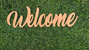 An outdoor scene showing a wooden welcome sign leaning against a stone wall surrounded by greenery