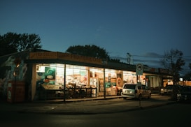 A small grocery store with large windows illuminated from the inside, displaying various colorful advertisements and products. The store exterior features a yellow awning with the name 'Fruiterie Cité' and is surrounded by parked vehicles. The scene is captured during dusk or early evening, creating a moody and quiet atmosphere.