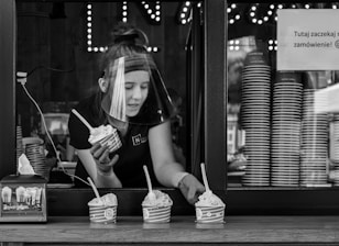 A person wearing a face shield is serving ice cream through a window. The window is lined with stacked cups, and three cups of ice cream with spoons are placed on the ledge. The setting suggests a casual food service environment.