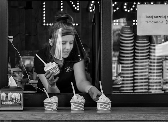 A person wearing a face shield is serving ice cream through a window. The window is lined with stacked cups, and three cups of ice cream with spoons are placed on the ledge. The setting suggests a casual food service environment.