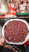 A person holds a large white sack filled with dried rose petals. The background includes a variety of items such as packaged sugar and large chunks of brown sugar or jaggery, all arranged on a colorful market stall.