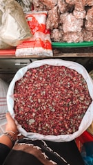 A person holds a large white sack filled with dried rose petals. The background includes a variety of items such as packaged sugar and large chunks of brown sugar or jaggery, all arranged on a colorful market stall.