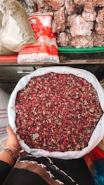 A person holds a large white sack filled with dried rose petals. The background includes a variety of items such as packaged sugar and large chunks of brown sugar or jaggery, all arranged on a colorful market stall.