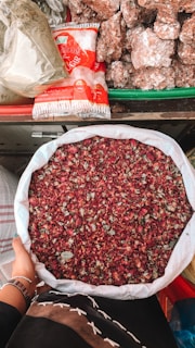 A person holds a large white sack filled with dried rose petals. The background includes a variety of items such as packaged sugar and large chunks of brown sugar or jaggery, all arranged on a colorful market stall.