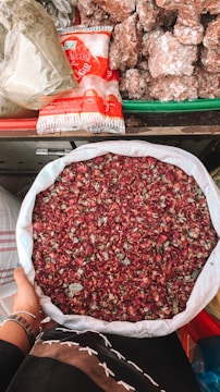 A person holds a large white sack filled with dried rose petals. The background includes a variety of items such as packaged sugar and large chunks of brown sugar or jaggery, all arranged on a colorful market stall.