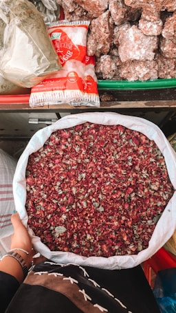 A person holds a large white sack filled with dried rose petals. The background includes a variety of items such as packaged sugar and large chunks of brown sugar or jaggery, all arranged on a colorful market stall.
