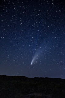 A clear night sky filled with stars, featuring a bright comet with a vivid tail extending across the sky. The silhouette of a landscape with gentle hills is visible at the bottom, emphasizing the vastness of the sky.